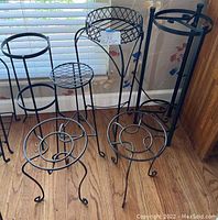 Photo showing all six black metal plant stands grouped together indoors on wooden floor near window.