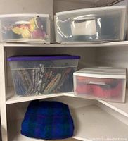 Plastic storage bins stacked in corner shelves displaying folded fabric in various colors and prints. One fabric folded piece with blue, purple, green plaid pattern set below bins.