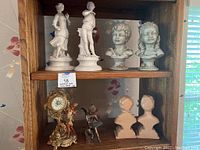 Overall view of two porcelain statues, two child busts, decorative clock with ornate base and figurines, brass cherub, and pair of bust bookends
