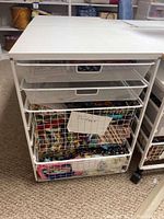 White Elfa storage unit with four metal mesh drawers labeled and filled with assorted fabric