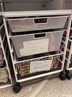 Front view of white metal Elfa storage cart with three mesh drawers on black caster wheels. The middle drawer has a paper label listing fabric types: Japanese, Fashion, Coffee, Tea; bottom drawer labeled French Rooster.