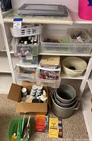Shelf with plastic storage bins holding fabric dye bottles, paints, and dyeing pots along with some paper and boxes.