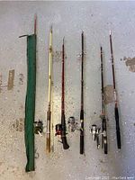 Five fishing rods laid flat on a concrete floor, four with reels and one without reel, along with a green canvas rod holder sleeve.