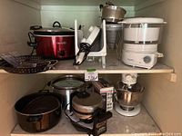 Kitchen shelf with multiple kitchen appliances and cookware including Crock-Pot, steamer, food grinder, waffle maker, pots, and sifters.