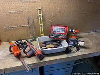 Workbench surface with bush trimmer, battery-powered chainsaw, hand saw, 24-inch level, power sander, battery-powered drill, red ratchet socket set case and a white bin with various hand tools.