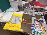 Photo showing painting clipboard easels, paint palettes with lids, tracing paper, color wheel, and paintbrushes in containers on a table