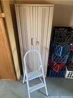 Closed view of beige plastic lockable cabinet with ribbed design and metal lock, alongside white two-step metal stool