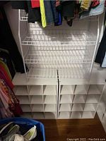 Photo showing five white cubical shoe bins side by side with 15 compartments each, placed on the floor in a closet with hanging clothes above.