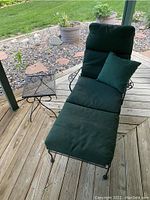 Photo showing black wrought iron adjustable lounge chair with green full-length cushion and a small matching pillow, alongside a small matching wrought iron end table with mesh top, all placed on wooden deck with greenery in the background.