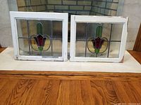 Front view of two stained glass window panels showing red tulip motif and white wood frames on wood surface in front of fireplace.