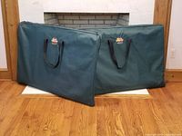 Two green fabric dog crates folded flat with black carrying handles, placed side by side on wooden floor.