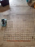 Three chrome gridwall panels lying flat on the floor, showing their size and rectangular grid pattern, with two wooden rulers for scale at the bottom.