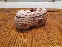 Photo of the ceramic peanut-shaped jar from a side angle on a wooden floor, showing the textured peanut design and brown nut details on the lid and body.