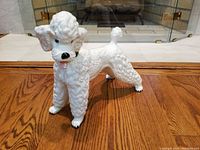 Front and three-quarter view of the white ceramic poodle dog figurine showing the textured fur, face details including black nose and pink tongue.