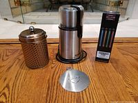 Full setup showing the KitchenAid electric coffee grinder, stainless steel bean storage vessel, a dusting stencil, and the box of light-up coffee stirrers on a hardwood floor.