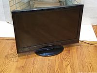 Front view of black Dynex 32 inch LCD TV on oval black pedestal stand, showing the screen and casing.