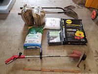 Various new garden supplies and used tools arranged on concrete floor, including bags of fertilizer, sand, soil mix, burlap bags, weed control fabric, mulch fork, shovel, and an electric weedwhacker.