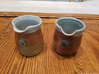 Front view of two handmade ceramic vessels with pour spouts, showing decorative emblems and rustic glaze.