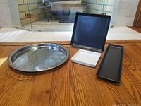 Four Ikea trays displayed on wooden floor including round silver metal tray, black matte square tray, black matte rectangular tray, and white gloss square tray.