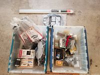 Two plastic bins on floor; first bin holds dryer vents, hose, digital timer switch, screws and other boxed hardware; second bin holds assorted nails, screws, hooks, wire, metal duct parts, small red hand tool, additional hardware bags.