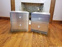 Front view of two silver brushed metal bedside tables with two drawers each and clear lucite drawer pulls, placed on wood floor in front of fireplace.