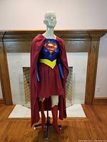 Front view of life-size Superwoman mannequin dressed in blue, red, and yellow costume with iconic 'S' emblem. Mounted on antique cast iron rolling base with wheels.