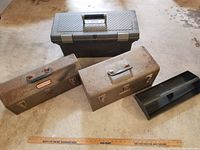 Three toolboxes: two metal vintage boxes and one modern plastic box, placed on a concrete floor next to a wooden ruler for scale.