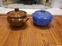 Front view of two covered stoneware casserole pots, brown left and blue right, showing lids and handles clearly