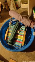 Photo of folded beach chair with multi-color striped fabric and wooden armrests inside blue plastic baby pool. The green camp stool and purple tent bag are visible folded on top.