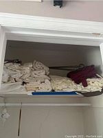 Multiple folded curtains in cream, burgundy, and brown colors stacked on a shelf, with visible curtain rods behind.