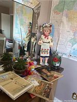 Photo showing doll in traditional costume, assorted books including Princess Diana and Princess Grace biographies, small potted plants on surface, with map of Brazil reflected in mirror