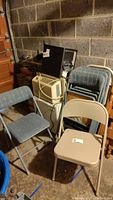 Shows four blue-gray fabric back and cushion folding chairs and two gray all metal folding chairs arranged near a wall with an air conditioning unit and other storage.