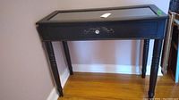 Front side view of black console table with glass top, turned legs, and decorative single drawer.