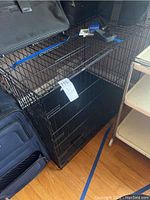 Left side angled view of black metal dog crate with plastic tray inside, tag 715 attached to door, shows scuffing and scratches.