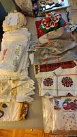 Photo showing assorted folded linens including lace-trimmed items, embroidered cloths with floral and fruit motifs, along with a colorful Christmas fabric piece.