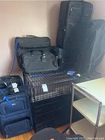 Eight pieces of mixed size black and blue soft-sided Hartman luggage including garment bag, duffels, and suitcases, stacked and grouped in room.