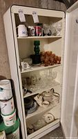 White metal cabinet filled with various mugs, glassware, plates, and serving ware on four shelves.