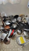 Photo showing various pots, pans, bundt cake pans, kettle, grill pan, wok, lids, and food service ware on floor, depicting items included in lot.