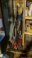 Image showing collection of multiple cleaning and gardening tools including brooms, shovels, and rakes stored upright in corner.