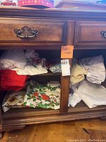 Table linens folded and stacked in a cabinet drawer under a wood dresser. Includes red, white, holiday themed and off-white linens with embroidered and printed holiday holly patterns.