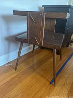 Two-shelf mid-century modern wooden table with unique decorative wood panel on the side, angled legs, some surface scuffs and scratches, placed on hardwood floor.