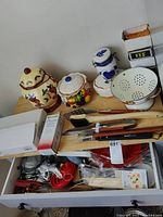 Various kitchen utensils, decorative ceramic jars, BBQ tools and white metal colander arranged on table and in drawer.