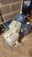 Two white rectangular 5-gallon jugs and two blue large water jugs with a smaller blue jug, all empty and placed on a concrete floor near a wall.