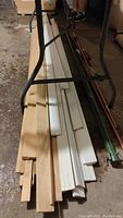 Photo showing assorted wood and trim pieces including natural wood and painted white pieces about 8 feet long, laying on the ground under a metal frame.