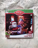 Front of the puzzle box showing Rudolph Red-Nosed Reindeer theme with Santa Claus and other characters, labeled as 100 piece puzzle for ages 3+. Box appears new and unopened.