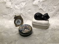 Photo showing front of mini brass photo frame shaped like cat head, round illustrated tin box, and porcelain trinket box with black cat figurine lid