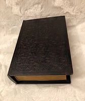 Top angled view of the closed book-shaped container showing detailed embossed pattern on cover and simulated gold pages.