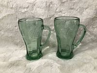 Pair of green Coca-Cola pressed glass mugs with handles shown upright on soft fabric background. Both have embossed logos and contoured thick glass design.