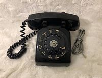 Front view of black rotary dial phone with coiled receiver cord and wall cord.