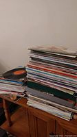 Large stack of vinyl records piled on a wooden table showing varying cover colors and sizes.